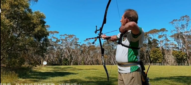 A photo of Garry with his bow stringed, pointing it at a target in the distance.